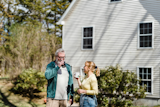 Older man and woman standing outside a white house, smiling and drinking from mugs on a sunny day.